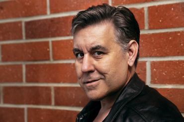 Man in black leather jacket posing against a brick wall with a confident smirk.