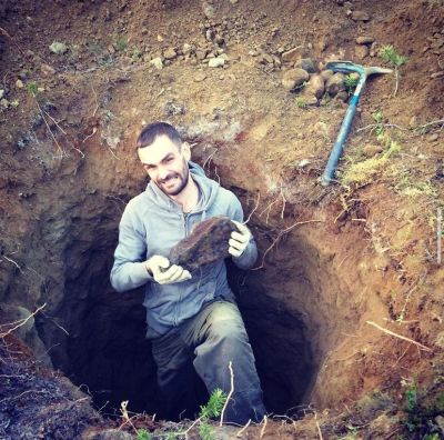 Man digging a deep hole holding a large rock with a pickaxe nearby.
