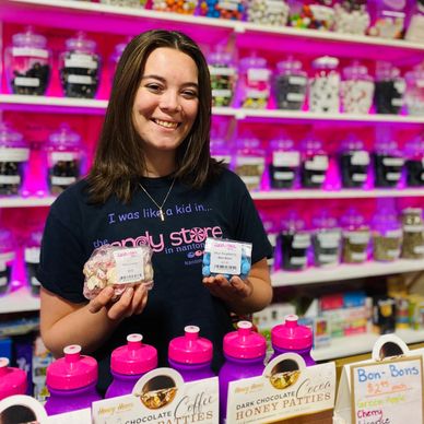 A smiling girl holding brightly coloured candies in front of a pink wall of candy jars