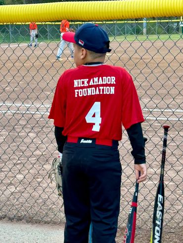 Youth baseball player in red jersey with number 4 stands by the fence holding a bat.