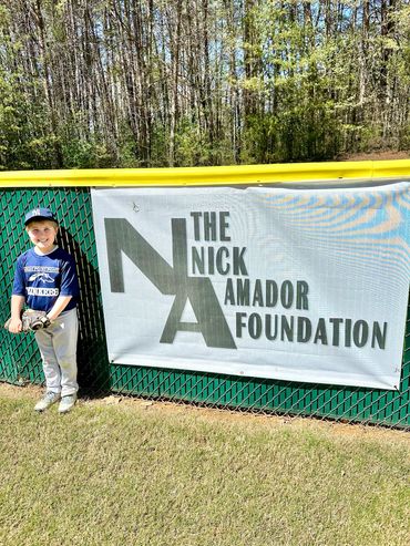 Young boy in baseball gear stands next to a Nick Amador Foundation banner at a baseball field.