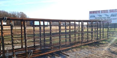 Rusty metal gates stacked outdoors near a Hawkeye Steel Sales sign.
