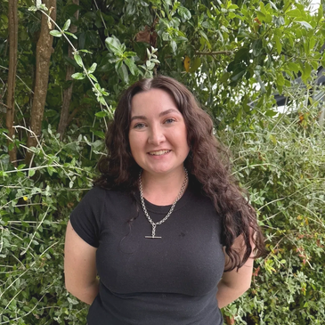 Smiling woman with long curly hair stands in front of lush green foliage.