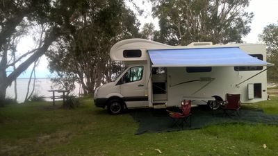 Camper van parked near lake with outdoor chairs and awning.