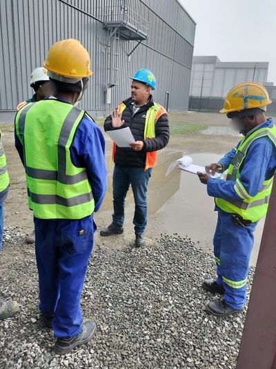Construction workers in safety gear having a site meeting outdoors near industrial buildings.