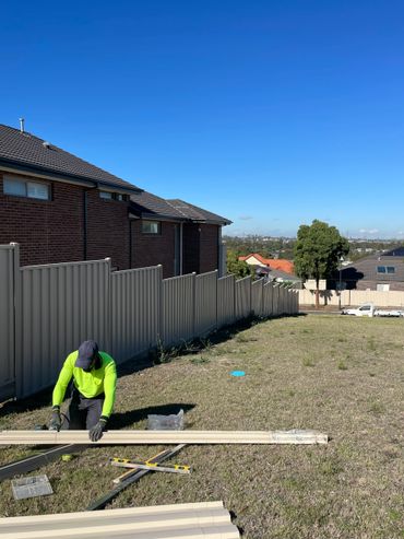 Worker cutting materials outdoors near a fenced residential area.