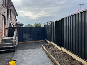 Backyard with black metal fences, brick house, and a yellow bucket on the ground.