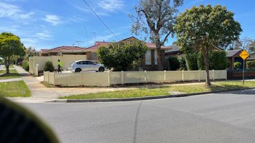 Suburban house with a white fence, trees, and a person walking near a silver car on a sunny day.