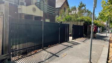 Two men talking near open black fence gates on a sunny sidewalk.