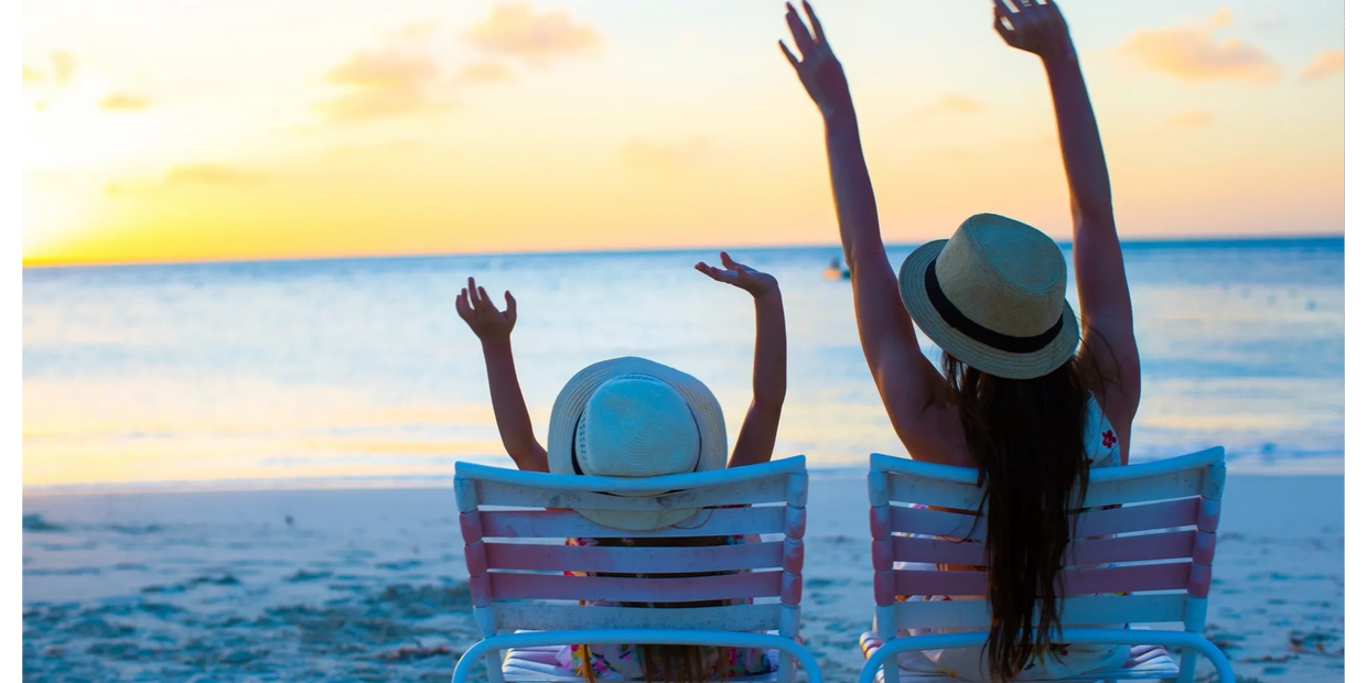 Mother and daughter arms stretched up high, sitting at the beach on chairs, sun set is colourful