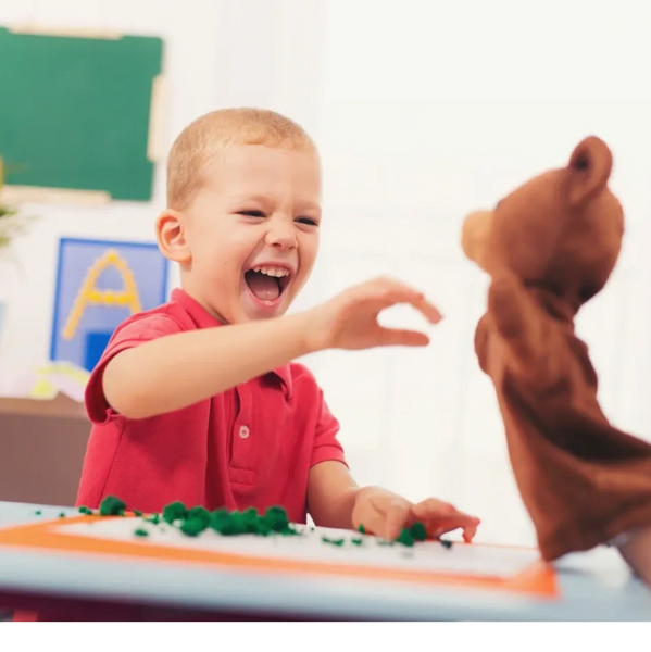 Young boy showing joy with a big smile, hands out towards a brown bear hand puppet