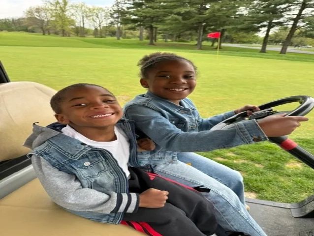 Two smiling kids in a golf cart on a green field.