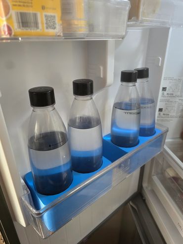 Four water bottles with black caps on a blue rack inside a refrigerator door.