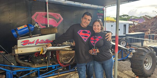 Couple wearing matching Superman shirts beside a dragster car.