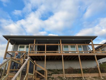 A man stands on wooden stairs leading to a house with a large porch under a blue sky.