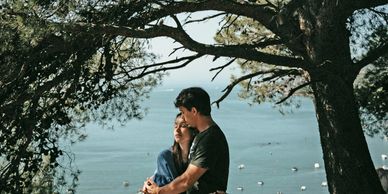 A couple hugging under a tree looking over water with boats