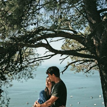 A couple hugging under a tree looking over water with boats