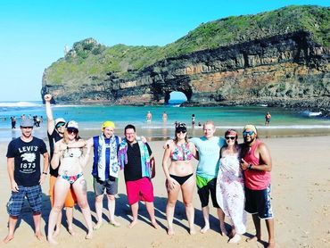 Group of friends posing on a sunny beach with a rocky arch formation in the background.