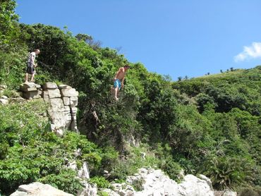 A man jumps off a rocky cliff into water below on a sunny day.