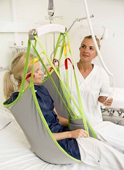 A woman in a human care overhead lift being helped from bed by a female caregiver
