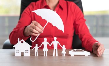 A person holds a paper umbrella over paper family, house, and car figures symbolizing protection.