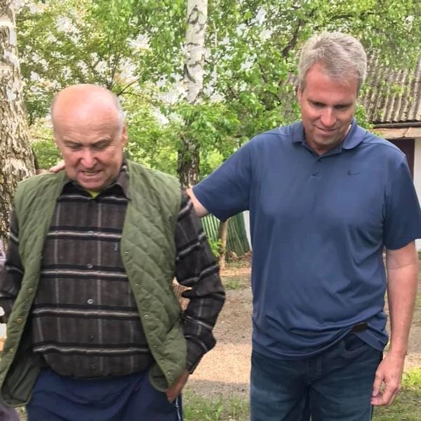 Two men walking outdoors, one supporting the other in a green vest and striped shirt.