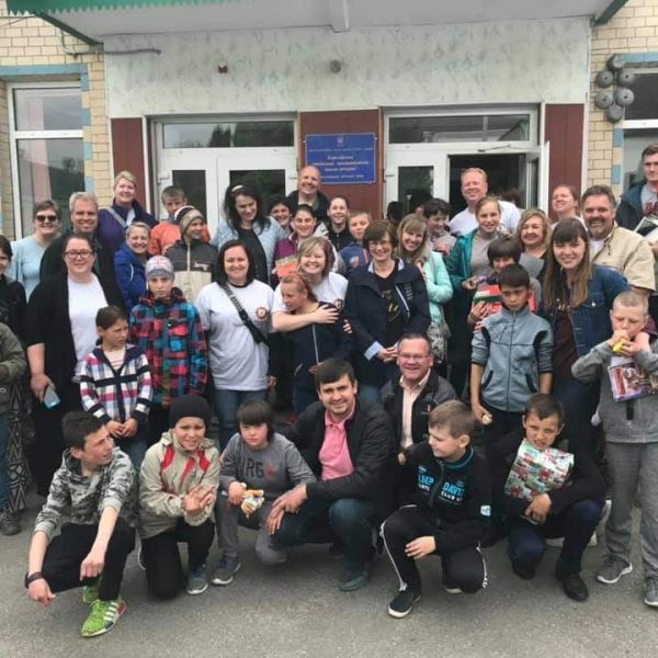Large group of smiling people, including children and adults, posing outside a building.