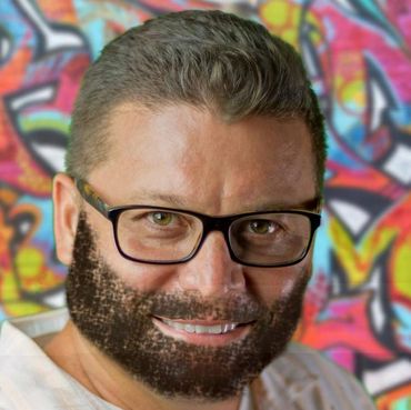 Man with glasses and a full beard in front of colorful graffiti.