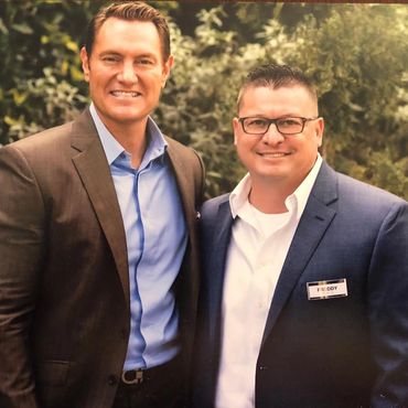 Two men in suits smiling outdoors with greenery in the background.