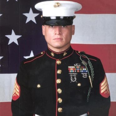 U.S. Marine in dress blues uniform standing in front of American flag.