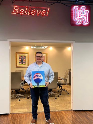 Man holding a RE/MAX hot air balloon photo in an office with a neon Believe sign.