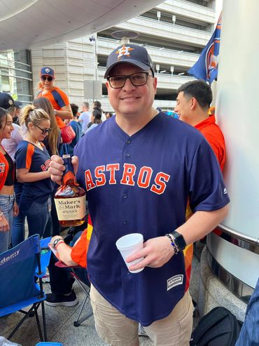 Man in Astros jersey holding a Maker's Mark whiskey bottle and cup at a crowded event.