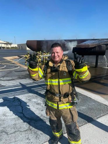Smiling firefighter in full gear posing with peace signs at a training site.