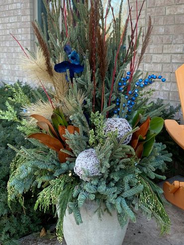 Outdoor pot decorated with winter greens, berries, magnolia, spruce tops, juniper, pine, and lights.