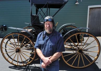 Man in vintage aviator gear stands in front of an old carriage.