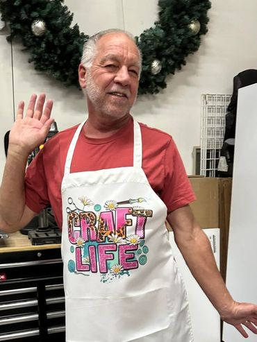 Smiling man waving, wearing a colorful "Craft Life" apron.