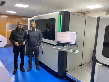 Two men standing beside a large digital printing machine in a clean industrial room.