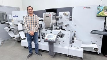 Man standing next to a large industrial printing machine in a factory setting.