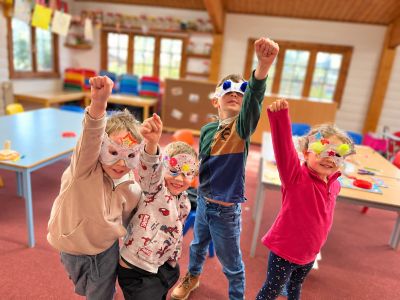 Children wearing colorful masks, raising their fists joyfully indoors.