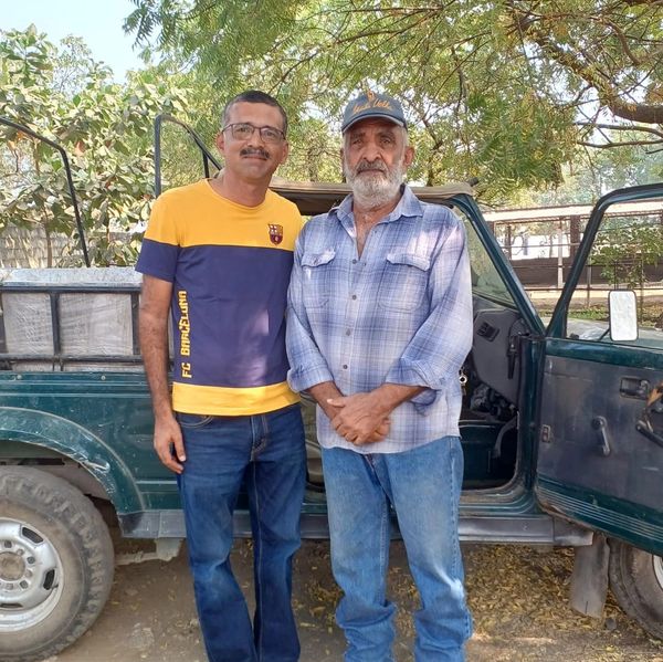 Two men standing beside a green vehicle with an open door under trees.