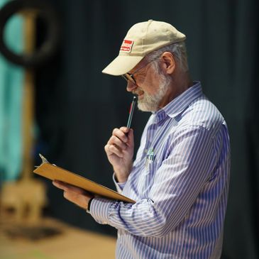 An elderly man in a cap and striped shirt examines something with a magnifying glass.