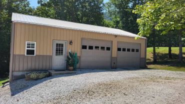A beige metal garage with two doors and a side entrance, surrounded by trees and gravel driveway.