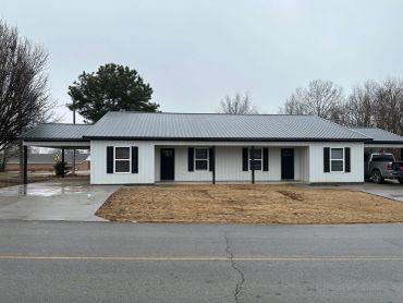 A simple, single-story house with a metal roof and black shutters on a cloudy day.