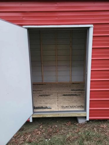 Open door of a red shed showing unfinished interior with plywood floor and wall studs.