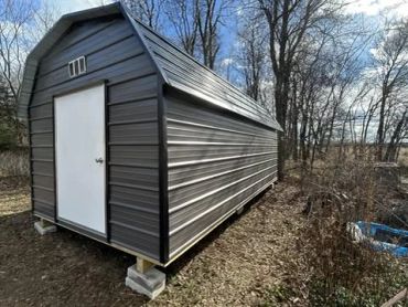 A black metal storage shed with a white door in a wooded area.