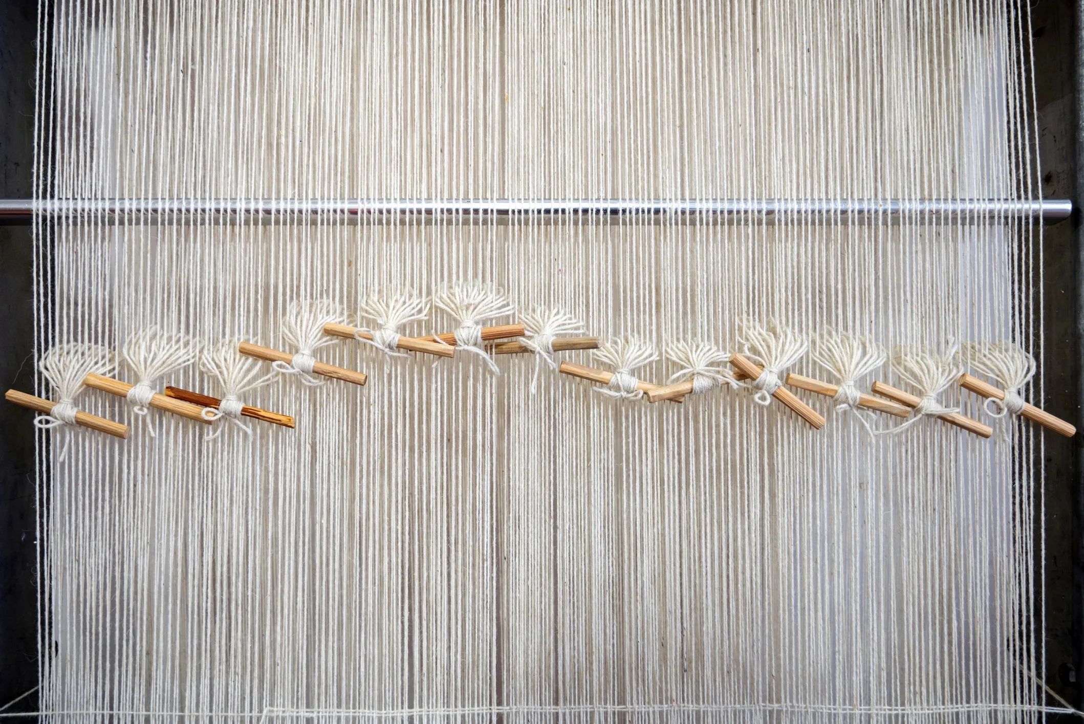 Close-up of white threads stretched on a loom with wooden sticks tied in patterns.
