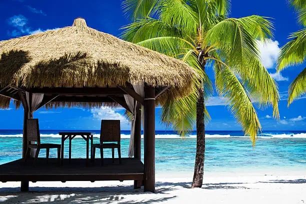 A thatched-roof beach hut with loungers and wooden chairs on a white-sand beach in the Cook Islands.