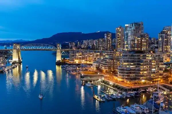 Night view of a city harbor with lit buildings and a bridge.