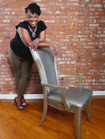 Smiling woman leaning on a silver chair against a brick wall.