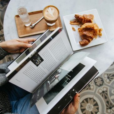 Person reading a book with coffee and croissants on a table.